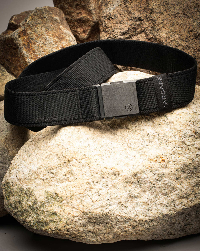 A black Arcade brand travel belt with stretch webbing is displayed coiled on light and dark rocks, highlighting its sturdy, modern design.