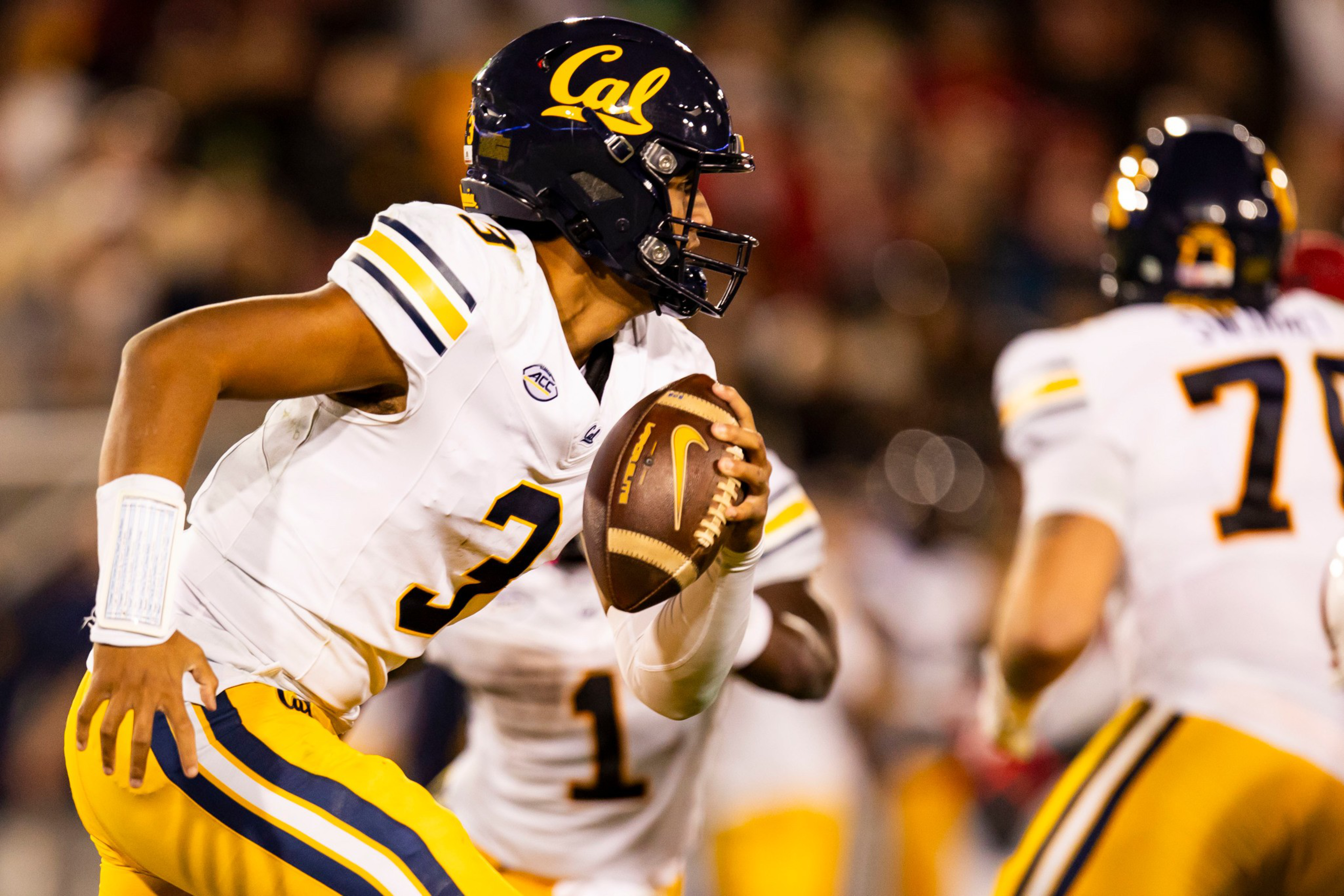 A football player wearing a white and yellow Cal uniform with number 3 runs while holding a football, with teammates in the background.