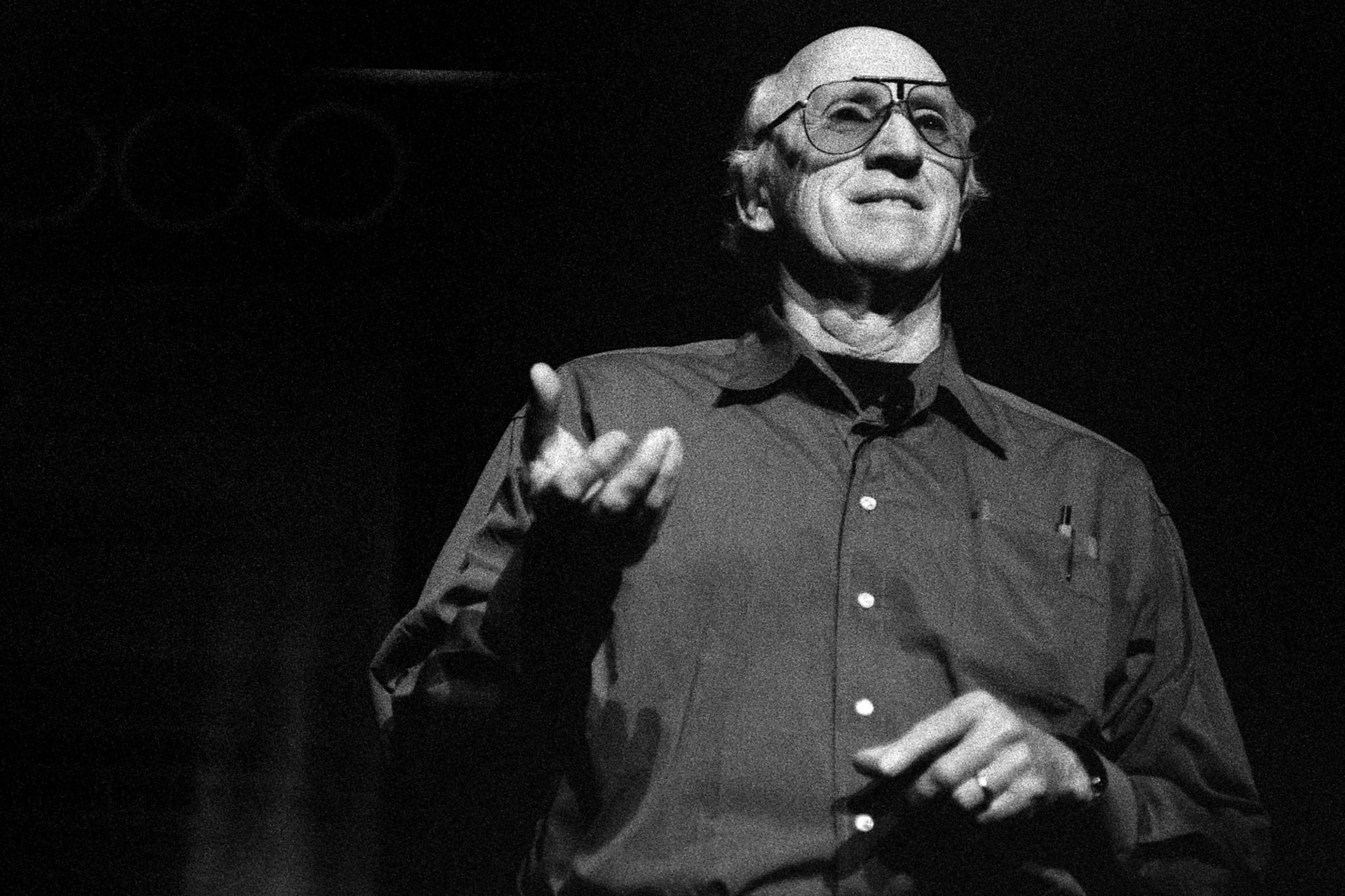 An older man wearing glasses and a buttoned shirt gestures with one hand while speaking on stage against a dark background.