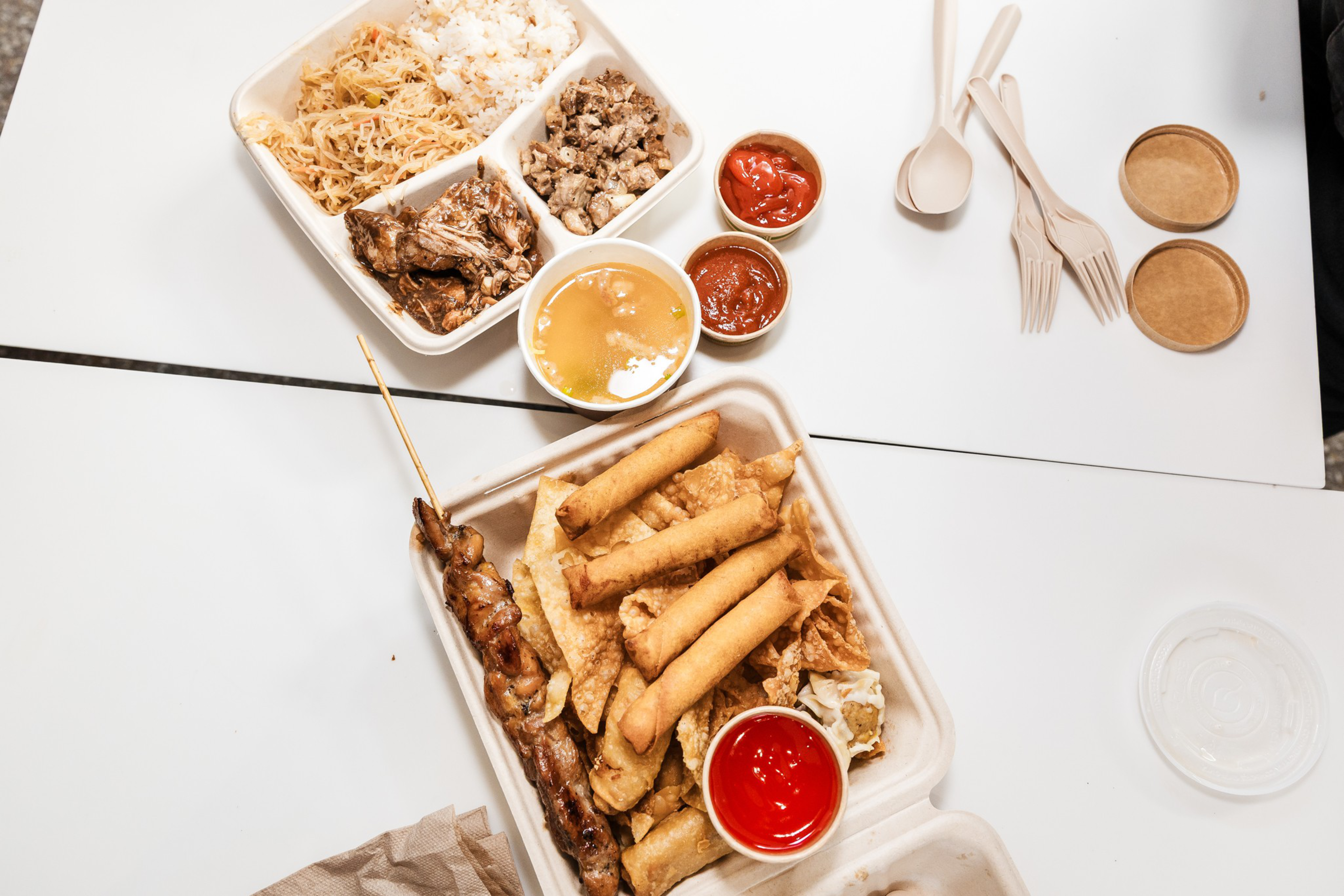 Two takeout containers with various fried foods, grilled meat skewers, shredded rice noodles, chopped meat, two types of dipping sauces, plastic forks, and spoons.