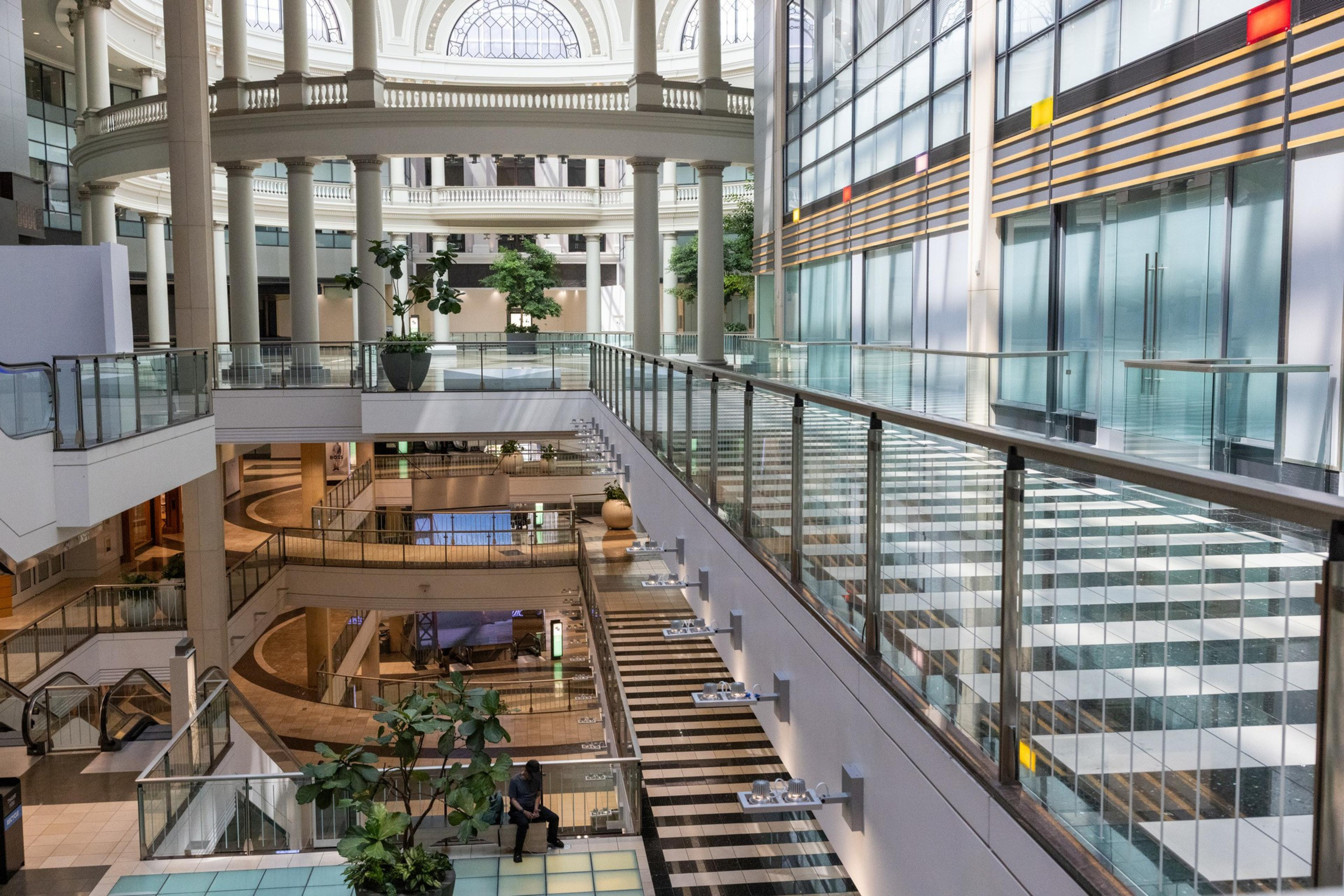 A spacious, multi-level indoor area with glass railings, large columns, plants, and natural light streaming through arched windows and a glass ceiling.
