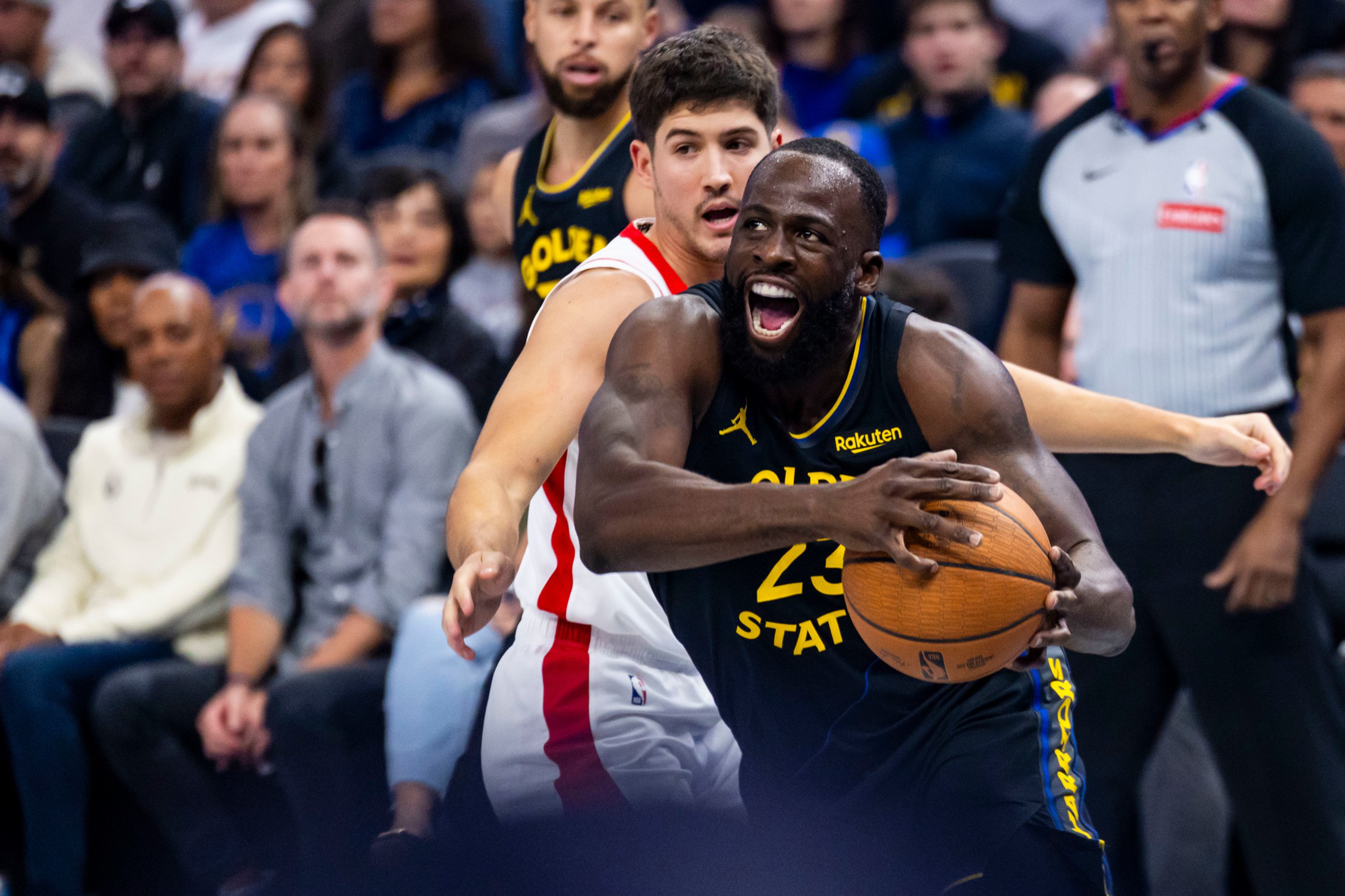 A basketball player in a dark Golden State Warriors jersey holds the ball tightly, guarded closely by a player in a white and red jersey.
