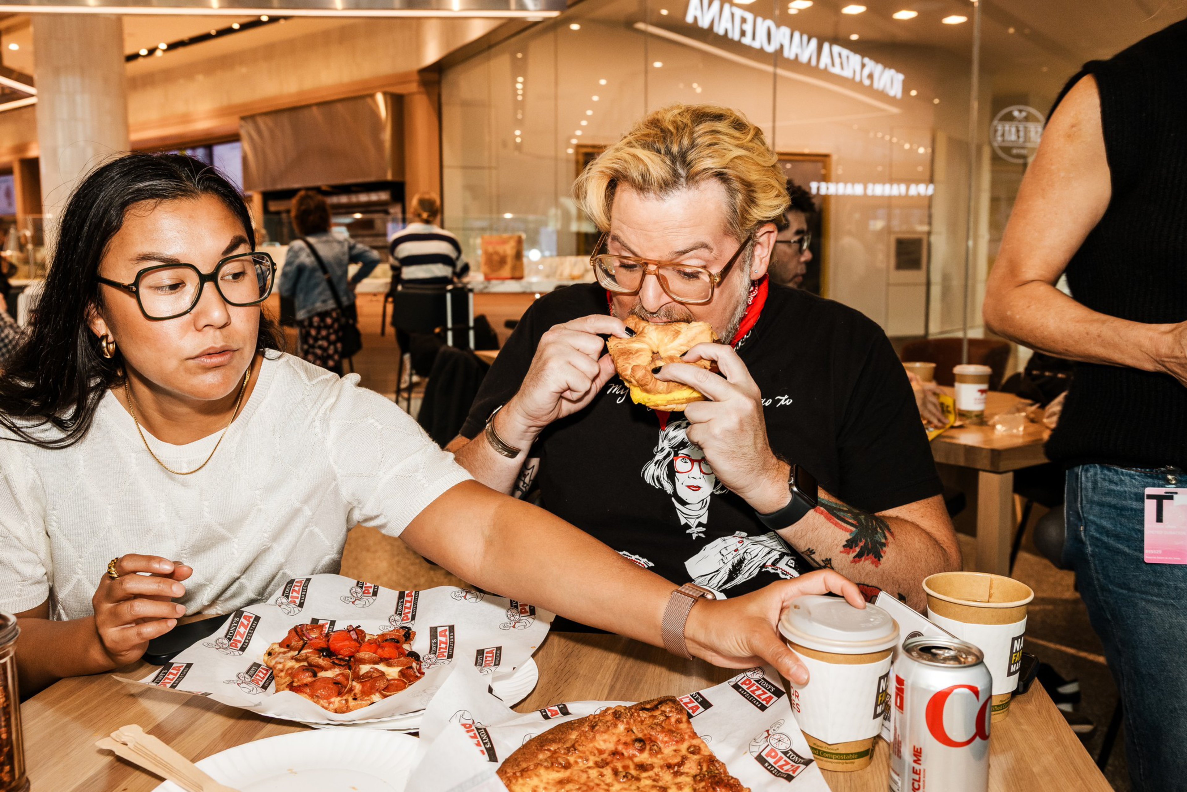 Two people sit at a table eating pizza, one woman reaching for a coffee cup while the man bites into a slice, with soda and pizza on the table.