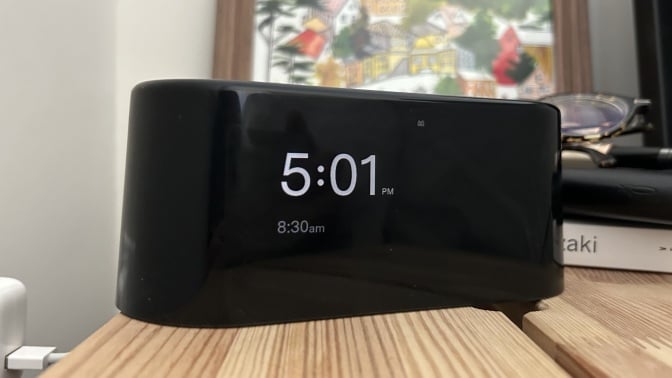 loftie alarm clock on nightstand in front of painting, pile of books, and glasses