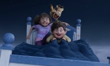 Two kids and a stuffed giraffe fly on a bed through the night sky.