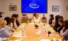 A group of teens sit at a long conference table and look toward a woman (Larissa May) speaking at the head. 