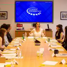 A group of teens sit at a long conference table and look toward a woman (Larissa May) speaking at the head. 