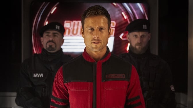 A man stands in a red jacket flanked by soldiers wearing black.