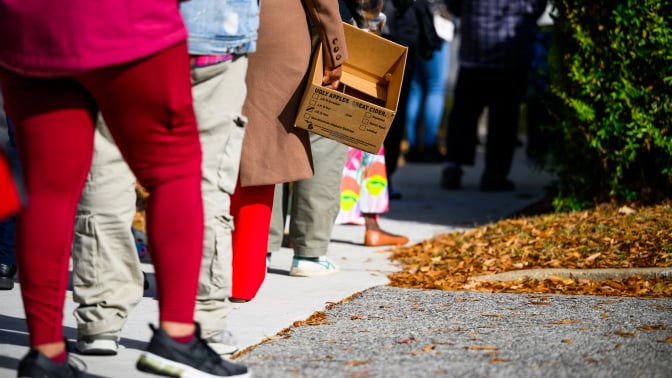 People stand in line holding cardboard boxes and grocery bags. 