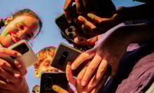 A group of young kids smile and hold out several different models of phones while huddled in a group.