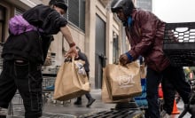 Two delivery drivers pass paper bags of groceries to each other on a city street.