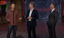 Three men stand on a talk show stage.