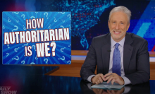 A man in a suit sits behind a talk show desk, grinning.