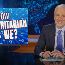 A man in a suit sits behind a talk show desk, grinning.