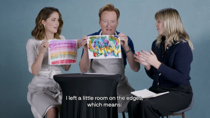 Three people sit behind a table covered with art supplies. Two of the people, a man and a woman, hold up paintings.