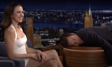 A woman laughs while sitting on a chair next to a man lying on a talk show desk.