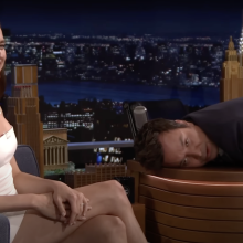 A woman laughs while sitting on a chair next to a man lying on a talk show desk.