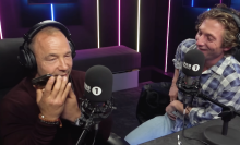 Two men sit in a radio studio wearing headphones and laughing, while one man speaks into a phone.
