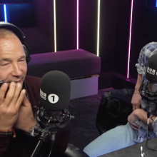 Two men sit in a radio studio wearing headphones and laughing, while one man speaks into a phone.