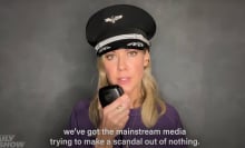 A woman in a pilot's hat stands against a grey wall, looking at the camera.