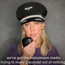 A woman in a pilot's hat stands against a grey wall, looking at the camera.