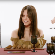 A woman sits at a table with wings in front of her and dark goblets.