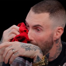 A man sits in a dark studio dabbing his eyes with a red napkin.