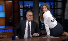 A woman wearing a "We Heart Stephen" shirt sits on a desk next to a man sitting down in a chair wearing a suit.