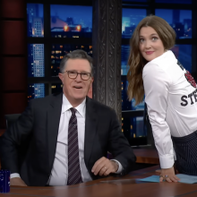 A woman wearing a "We Heart Stephen" shirt sits on a desk next to a man sitting down in a chair wearing a suit.