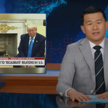 A man in a suit sits behind a talk show desk. In the top-left is an image of two other men in suits in the Oval office.