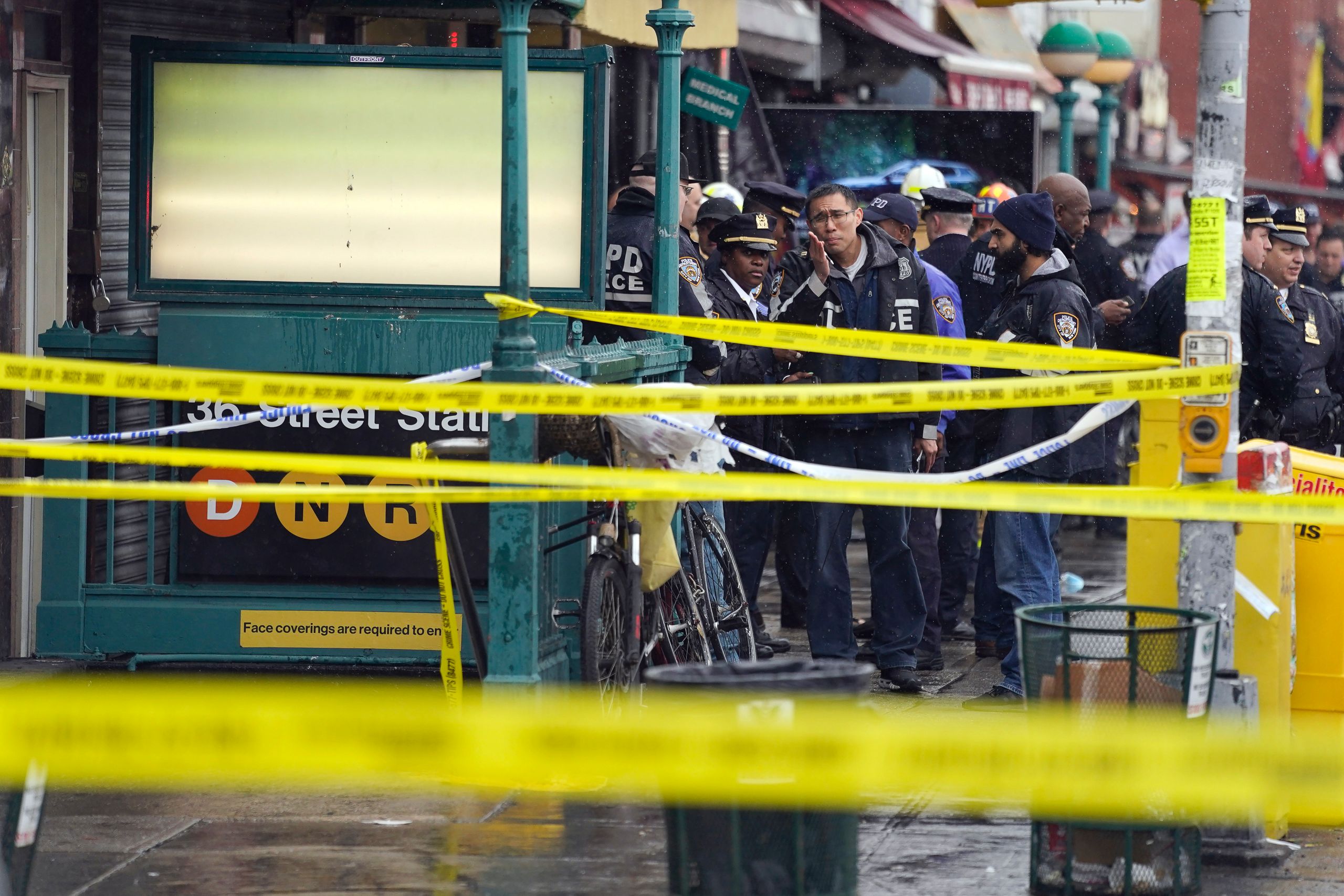 Yellow caution tape cordoning off the 36th Street Subway station entrance. Police officers stand on the sidewalk behind...